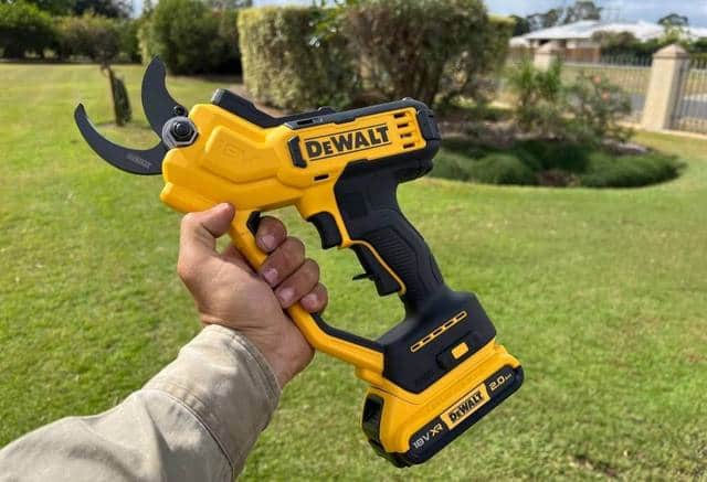 A hand holding a battery pruning tool in front of a lawn
